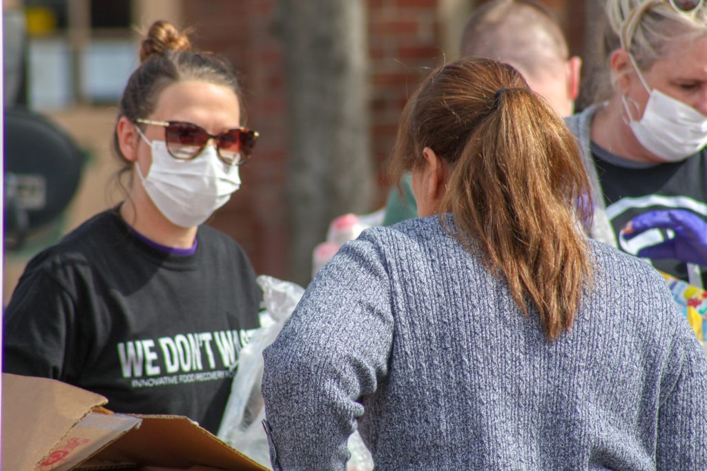 A woman wearing a white mask, sunglasses, and a black we don't waste teeshirt faces the camera. She is interacting with another person who's back is to the camera. They have long brown hair in a ponytail and are wearing a blue sweater.